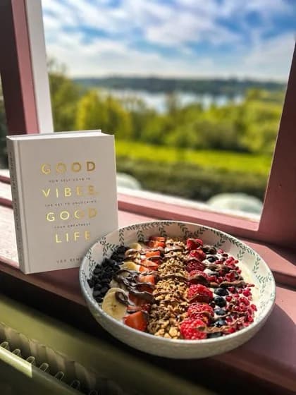 A Glow Bowl topped with fruit, granola and chocolate beside a countryside window.